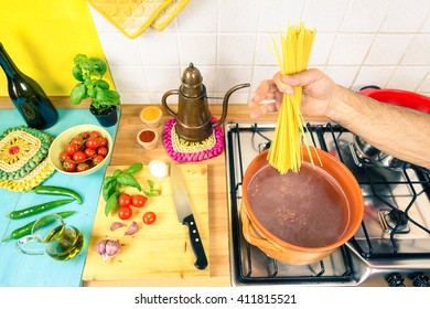 Man Hand Putting Down Spaghetti In Rustic Pot At Home Kitchen - Cook Holding Pasta Top View With Cookery Stove And Fresh Vegetables On Blue Wooden Board - Concept Of Healthy Traditional Food Cooking