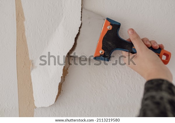 Man hand tearing off old wallpaper with scraper from wall.