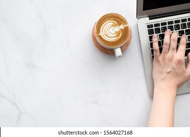 Man hand is typing on laptop computer keyboard over white marble office desk table with a cup of latte coffee. Top view with copy space, flat lay.