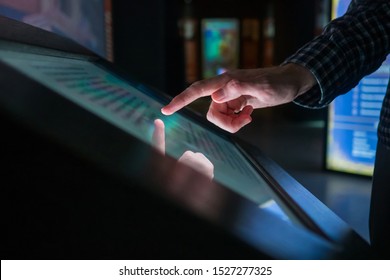 Man hand using interactive touchscreen display of electronic multimedia terminal at modern museum or exhibition. Evening time, low light illumination. Education, futuristic and technology concept