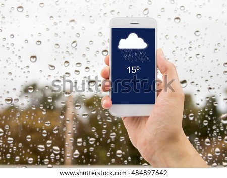 man hand using mobile smart phone with glass window full of water droplets of raining day view with weather forecast widget mobile application program template.