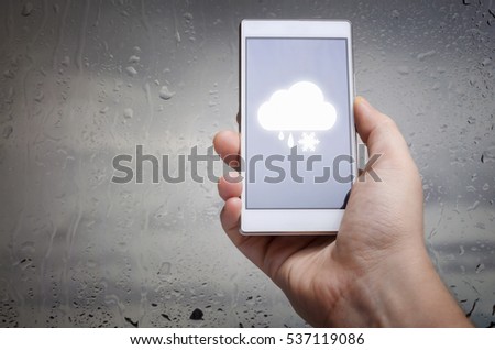 Man hand using mobile smart phone with glass window full of water droplets of raining day view with weather forecast widget mobile application program template