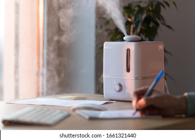 Man hand writes with pen in notebook near the aroma oil diffuser on the table, steam from humidifier, selective focus. Humidification of air in the apartment during the period of self-isolation 