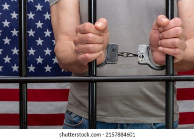 A man is handcuffed in a cell behind bars against the background of an American flag. Concept: the prisoner in the courtroom, the verdict of the court to the convicted person, prison term, law. - Powered by Shutterstock