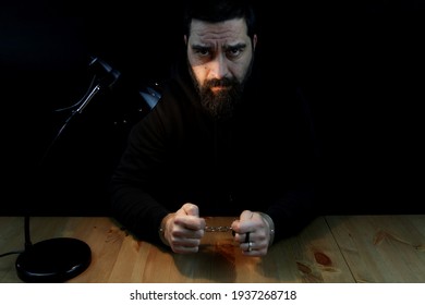 Man With Handcuffs, Arrested. Sitting Behind The Desk Looking At The Camera On Black Background. Police Interrogation Room.