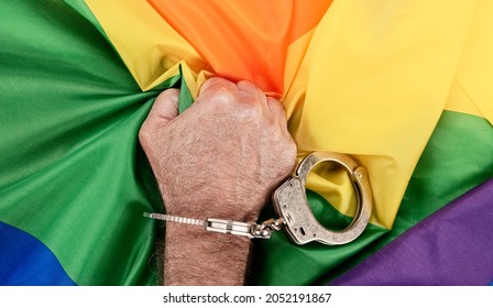 A man in handcuffs grasps the LGBTQ rainbow flag in frustration and anger as oppression and bureaucracy hinder acceptance - Shutterstock ID 2052191867