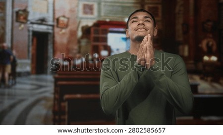 Man with hands clasped in prayer inside ornate church nave, smiling gently in green shirt; reverence faith.