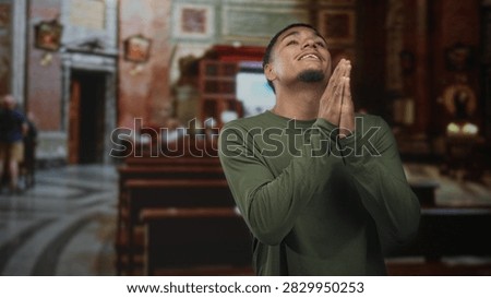 Man with hands clasped in prayer inside ornate church nave, smiling gently in green shirt; reverence faith.