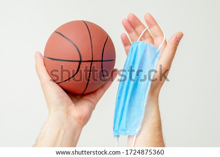 Man hands holding basketball ball with medical protective mask on light background during coronavirus. Basketball paused because coronavirus.