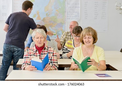 A Man Hands Out Books To A Classroom Of Older Adults.