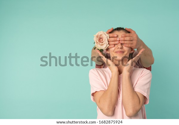 Man hands with rose covered\
eyes of his girlfriend, preparing surprise for 8 march, woman\'s\
day, mother\'s day, isolated over blue background with\
copyspace