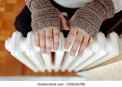 A man hands in wool gloves warm near the heater. Old men's hands in knitted gloves on heating radiator at home during the day. Person heating their hands at home over a domestic radiator in winter. - Shutterstock ID 2106326786