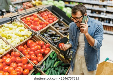 Man happily selects fresh tomatoes while chatting on his phone in a supermarket filled with colorful vegetables. This image captures a moment of healthy choices and balanced diet - Powered by Shutterstock