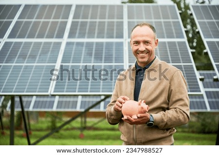 Man happy about his investments in alternative energy. Enthusiastic investor glad about earnings he got. Man with piggy bank in hands smiling to camera.
