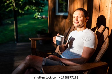 Man happy sitting relaxing on rocking chair lounge on porch of house in morning wooden cabin cottage drinking coffee or tea from cup mug - Powered by Shutterstock