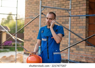 A Man In A Hard Hat And Construction Overalls Is Talking On The Phone At A Construction Site. High Quality Photo