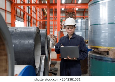 A man in a hardhat and mechanic coverall dust protection suit checks laptop computer within the metal sheet factory. Steel sheet roll stack are in the working area. - Powered by Shutterstock