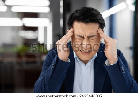 A man has a headache at the workplace inside the office, an Asian businessman in a business suit is sick, has a severe migraine, the boss is sitting at a desk with a laptop.
