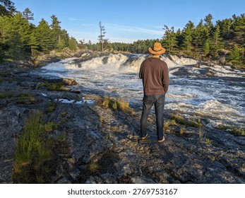 A man in hat admiring the view while standing on the rocky riverbank by a large rushing waterfall during sunset in the Canadian wilderness.