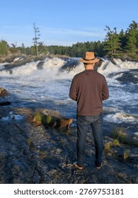A man in hat admiring the view while standing on the rocky riverbank by a large rushing waterfall during sunset in the Canadian wilderness.