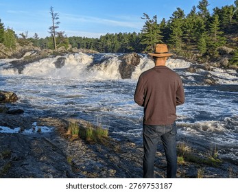 A man in hat admiring the view while standing on the rocky riverbank by a large rushing waterfall during sunset in the Canadian wilderness.