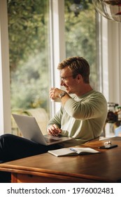 Man having coffee while working from home. Man sitting at table with laptop drinking coffee looking away and smiling.
