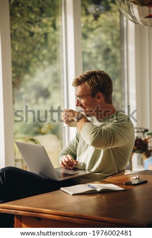 Man having coffee while working from home. Man sitting at table with laptop drinking coffee looking away and smiling.