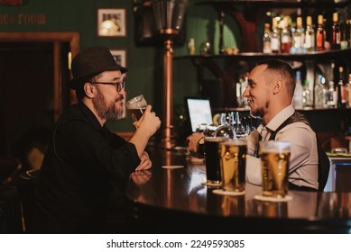 man is having fun chatting with a bartender in bar counter while drinking beer from a glass in a pub - Powered by Shutterstock