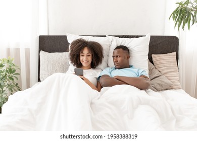 Man Having Problem While Girlfriend Using Smartphone In Bed. Shocked Young African American Guy Looking At Smiling Woman Mobile Phone, Husband Is Angry At Wife For Constantly Using Phone In Bedroom