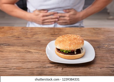 Man Having Stomach Pain Near Fresh Burger On Plate Over Wooden Desk