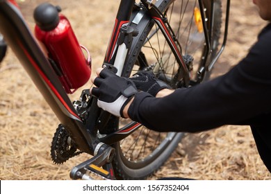 Man having stop, being outdoors, fixing bicycle by own forces, changing details, problems with movement, red bottle linked with bicycle, man wearing black clothes having problems. Bike repair.