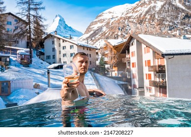 Man Having Wine While Swimming In Pool Against Town And Mountains. Tourist Enjoying In Hot Tub Against Matterhorn. Scenic View Of Snow Covered Landscape In Alps.