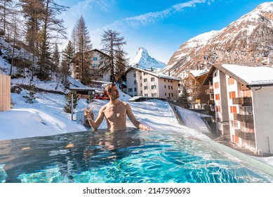 Man Having Wine While Swimming In Pool. Tourist Relaxing In Hot Tub Against Matterhorn Mountain And Houses. Scenic View Of Snow Covered Landscape In Alpine Region.