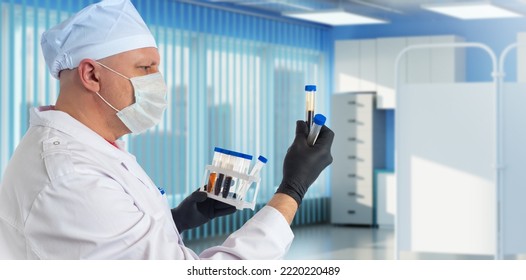 Man Is Head Doctor Of Clinic. Hospital Employee In Medical Mask. Head Doctor Is Holding Plastic Test Tubes. Man With Plastic Test Tubes In Interior Of Clinic. Head Doctor During Work.