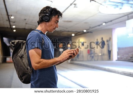 Man in headphones and backpack using smartphone while waiting in subway station