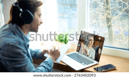 Man with headphones taking notes during online coaching video call at home. Concept of remote learning, client planning, business coaching, and online consultation.