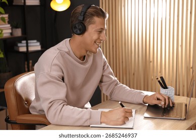 Man in headphones taking notes while passing online test via laptop at table indoors