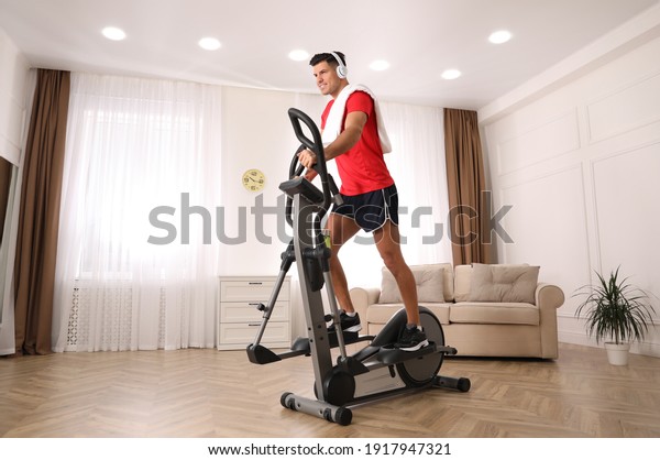 Man with headphones and towel using modern\
elliptical machine at home