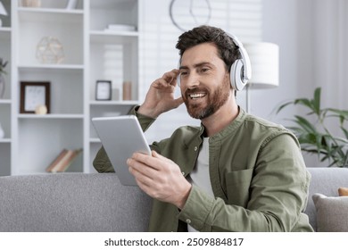 Man with headphones using tablet while relaxing on couch. Smiling, enjoying music or podcast in bright living room. Casual home environment promotes comfort and leisure.