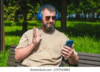 Man in headphones waves while using smartphone for video call on park bench. Concept of remote communication, online meeting, mobile lifestyle.