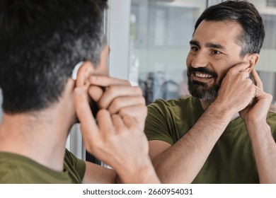 Man with hearing problems tests out his new behind-the-ear hearing aid, smiling as he observes himself in home mirror. Audiology
