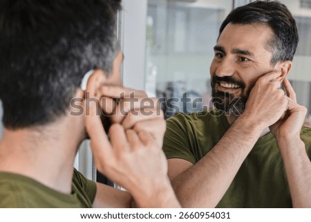 Man with hearing problems tests out his new behind-the-ear hearing aid, smiling as he observes himself in home mirror. Audiology
