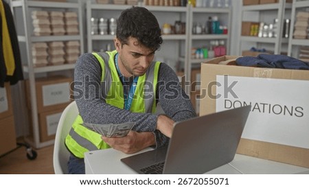 Man held cash while counting records in a warehouse by a hispanic volunteer beside a donation box near a laptop tracking money.