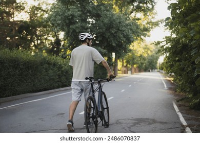 a man in a helmet walks next to a bicycle on a brick path, with a white railing and green grass to the sides. The sun is setting behind him, casting a warm glow over the scene - Powered by Shutterstock