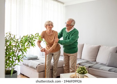 Man Helping An Elderly Woman Having A Back Pain. Sad Retired Woman With Walking Cane In Living Room Near Senior Husband. Senior Couple Looking At Each Other And Holding Hands At Home