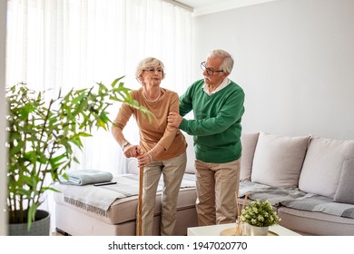 Man Helping An Elderly Woman Having A Back Pain. Sad Retired Woman With Walking Cane In Living Room Near Senior Husband. Senior Couple Looking At Each Other And Holding Hands At Home