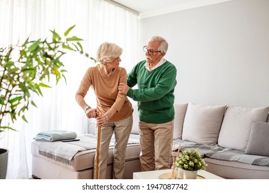 Man Helping An Elderly Woman Having A Back Pain. Sad Retired Woman With Walking Cane In Living Room Near Senior Husband. Senior Couple Looking At Each Other And Holding Hands At Home