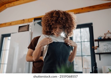 Man helping his smiling girlfriend or wife with curly hair to put an apron on, preparing to cook together in modern kitchen at home - Powered by Shutterstock