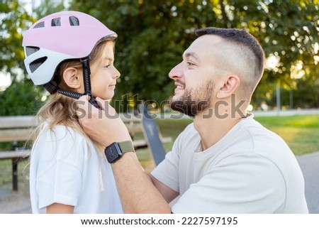 Man helps child girl fastens protective helmet for learning to ride skateboard at park. Father helping his daughter to wear a cycling helmet.