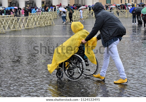 A man helps a person in a wheelchair\
to reach his destination by walking in the\
rain